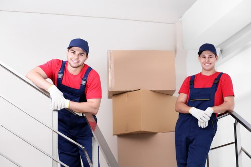 The image depicts two male removals workers dressed in red shirts, blue caps, and navy overalls with white gloves, standing inside a residential property in a staircase area. They are positioned on either side of a modern metal railing, smiling at the camera. Behind them, there are three large cardboard boxes stacked on top of each other, resting against a plain white wall that suggests a house or apartment in the Acton area. The staircase has a white-painted wall and a sleek, metallic handrail, indicating a contemporary interior environment typical of London suburbs or residential streets near Acton. The workers appear to be preparing for or engaged in a house removal, with the focus on their professional attire and the visible household items. The scene emphasizes careful handling of personal belongings during a local move, capturing the practical and organized nature of house removals services offered by companies like Acton Man with Van in West London.