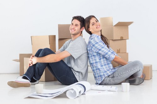 A young man and woman are sitting back-to-back on a clean white floor in a bright room, surrounded by multiple cardboard moving boxes of various sizes. The man, dressed in a light grey t-shirt and dark trousers, is smiling and looking slightly to the left, while the woman, wearing a blue checkered shirt and grey jeans, is also smiling and looking upward. They appear relaxed amidst the packing materials, with a few rolls of packing paper and two white mugs placed in front of them. The scene suggests a recent or ongoing house removal, with the boxes ready for transport. The setting features minimal decor, emphasizing the focus on the moving process, and the well-lit environment contributes to a positive, organized atmosphere associated with professional removals in the Acton area, London. Acton Man with Van offers household moving services, and the image captures the beginning stages of a typical domestic relocation, illustrating a friendly and efficient removal process in a residential context consistent with services near W3 postcode.