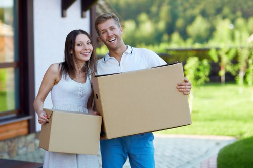 A young couple, appearing happy and smiling, stands outdoors on a residential driveway with a lawn and trees in the background, each holding a cardboard box used for house removals. The woman, on the left, has long dark hair and is dressed in a sleeveless, light-colored dress, while the man, on the right, has short blond hair and is wearing a light blue polo shirt with matching blue trousers. They are positioned in front of a modern house with a white exterior and wooden window frames, suggesting a suburban setting in Acton. The couple is engaged in the process of moving house, with the boxes likely containing personal belongings. The bright, natural sunlight illuminates the scene, emphasizing the cheerful atmosphere of the relocation activity. This image visually supports professional house removal services provided by Acton Man with Van, highlighting the friendly and efficient approach to local removals in the Acton area, located within postcode W3, London.