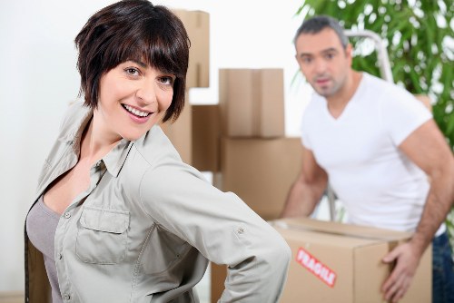 The image shows a woman with short dark hair and light skin, smiling and positioned in the foreground, wearing a light grey jacket over a grey top. She is in an indoor residential setting, possibly a living room or hallway, with moving boxes and cardboard packaging visible behind her. The background includes a man with short dark hair, light skin, and a beard, dressed in a white t-shirt, holding a medium-sized cardboard box sealed with red tape. The room features a green leafy plant and neutral-toned walls, suggesting a well-lit, tidy environment. The scene captures a moment during a house move in Acton, with the woman appearing confident and helpful while the man is preparing to load the boxes, aligning with the services offered by Acton Man with Van for local house removals. Few objects such as cardboard boxes, which vary in size and are packed with household items, are stacked or being carried, emphasizing the packing and moving process typical of professional removals in the area.
